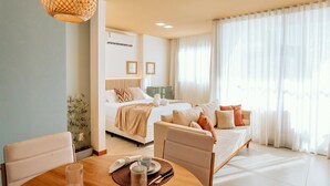Quarto de hotel moderno com cama, sofá, mesa de jantar e uma janela grande com cortinas leves.