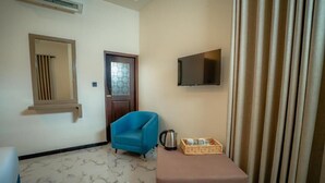 A hotel room with a blue armchair, a television mounted on the wall, a door with a frosted glass panel, and a small table with a basket.