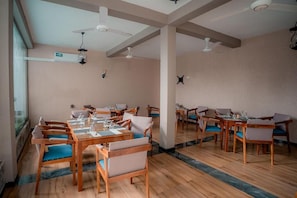 Uma área de refeições com mesas e cadeiras de madeira, ventiladores de teto e uma grande janela.