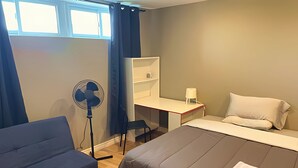 A room with a bed, a desk, a chair, a standing fan, and a window with curtains. - Basement Queen Bedroom-E（Private Room，Shared Bathroom） (Greater Sudbury)