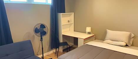 A room with a bed, a desk, a chair, a standing fan, and a window with curtains.
