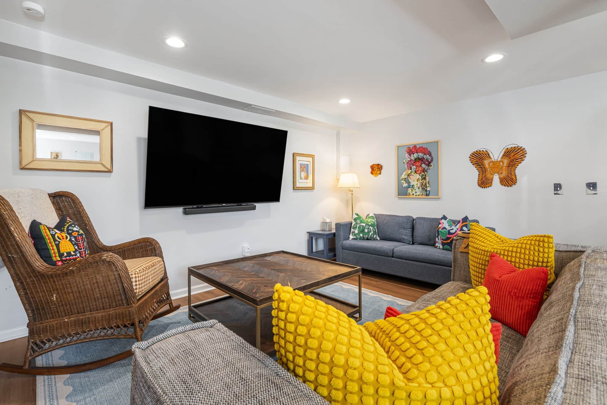 Una sala de estar con televisor de pantalla plana, muebles de mimbre, un sofá gris y cojines coloridos.