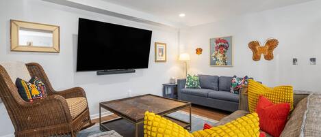 Una sala de estar con televisor de pantalla plana, muebles de mimbre, un sofá gris y cojines coloridos.