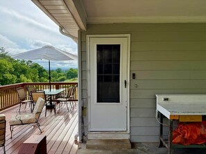 Um alpendre coberto com uma mesa e cadeiras, uma porta com janela, e vista para árvores e montanhas.