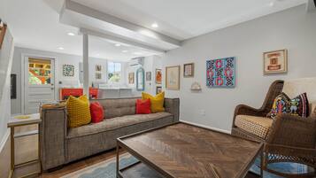 Ein modernes Wohnzimmer mit einer Couch, einem Couchtisch und einem Korbstuhl. An den Wänden befinden sich Kunstwerke, und ein Fenster sorgt für natürliches Licht.