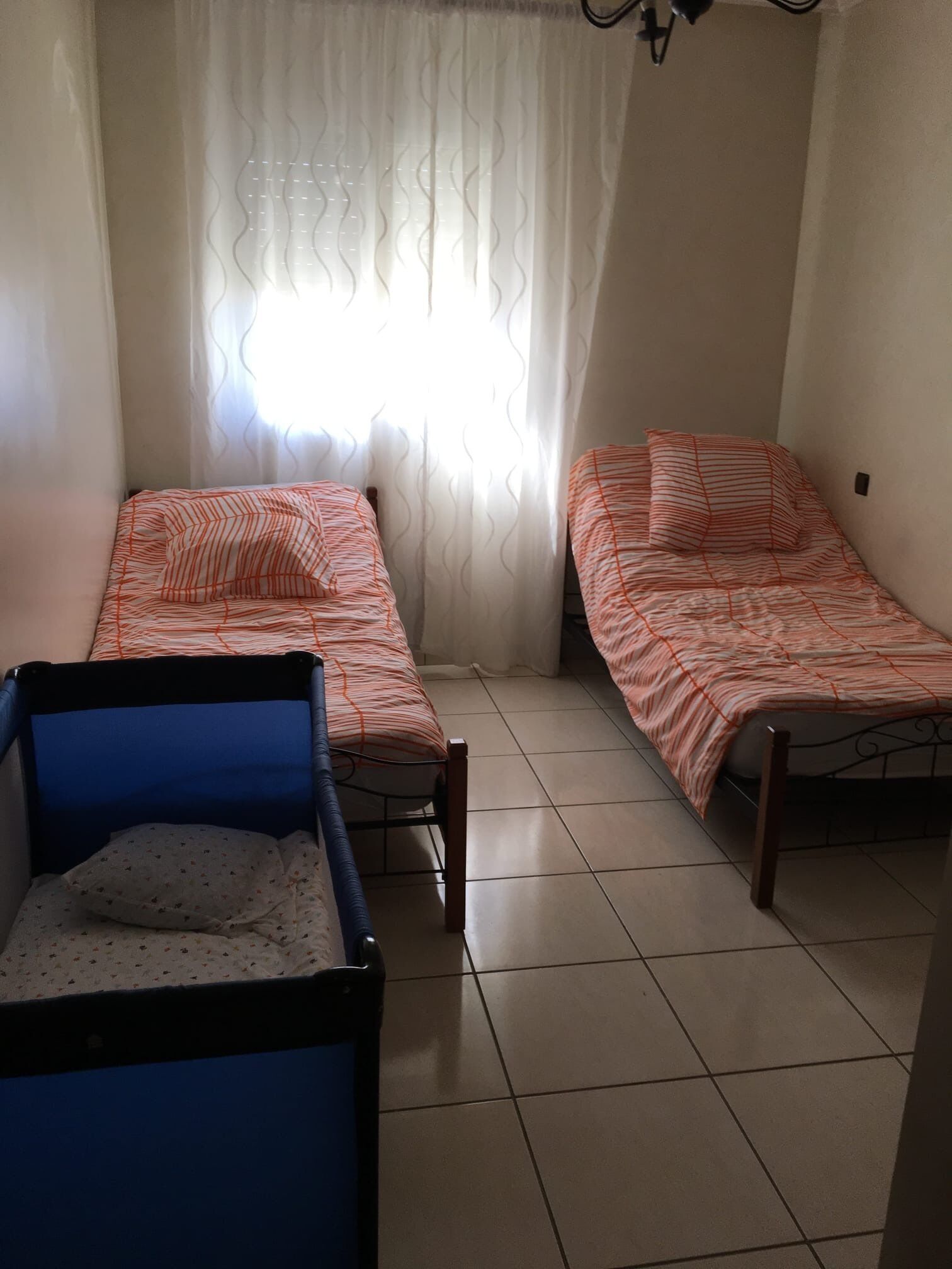 Quarto pequeno e bem iluminado com duas camas, uma cadeira azul e uma janela com cortinas leves.