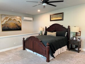 A bedroom with a wooden bed, a ceiling fan, an air conditioner, a framed painting, and a bedside table with a lamp.
