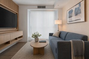 Ein modernes Wohnzimmer mit einem Flachbildfernseher, einem grauen Sofa, einem Couchtisch mit Pflanze und einem großen Fenster mit Jalousien.