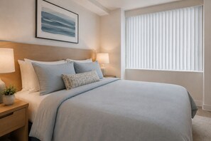 Ein ordentlich bezogenes Bett mit Kopfteil, zwei Nachttische mit Lampen, ein gerahmtes Kunstwerk an der Wand und ein Fenster mit Jalousien.