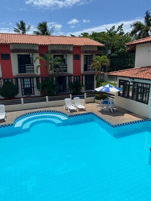 A swimming pool with lounge chairs and an umbrella, surrounded by buildings with red-tiled roofs and palm trees. - Nazareth Residential, apartment 16 (Porto Seguro)