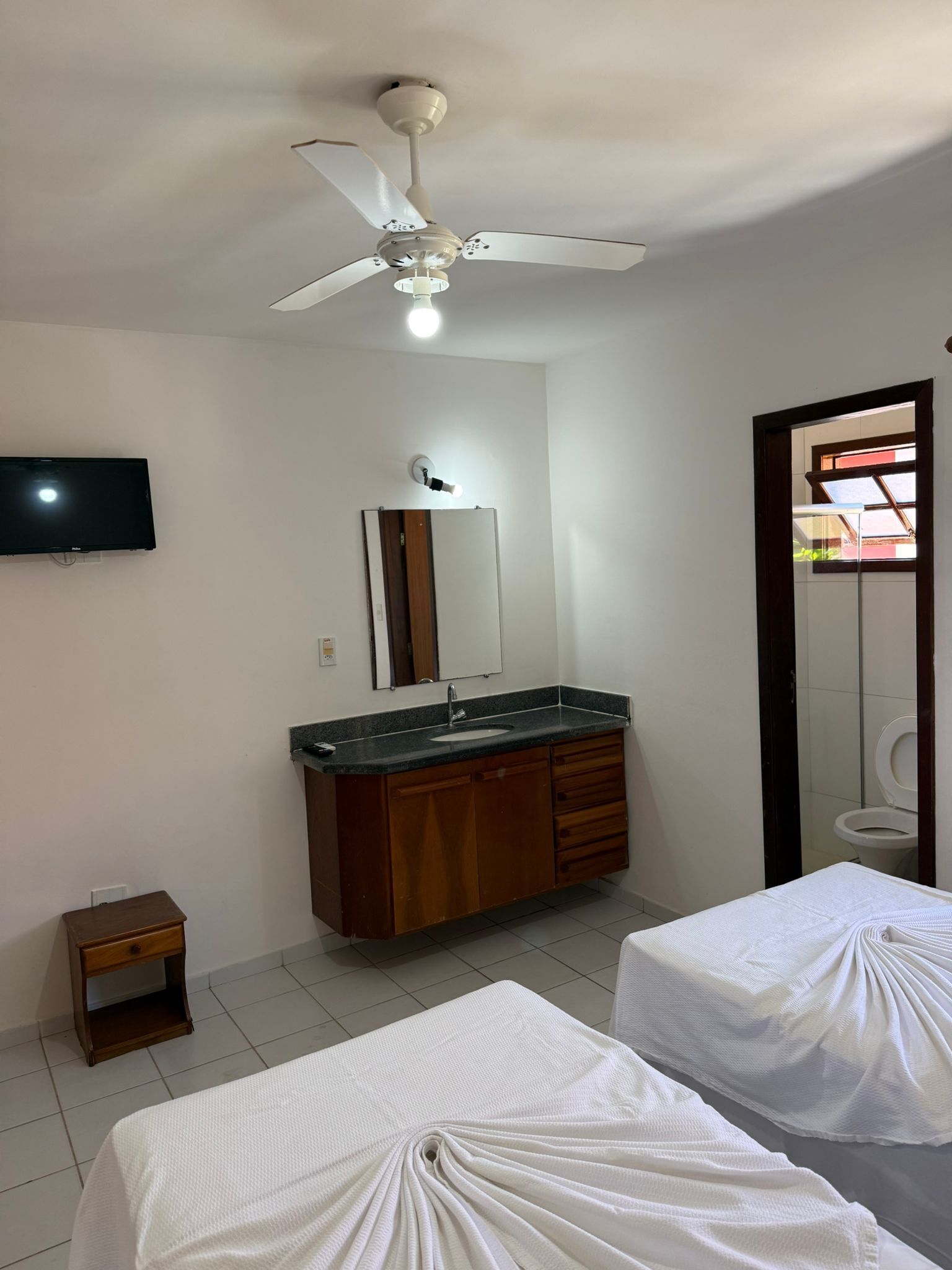 Un dormitorio con ventilador de techo, televisión, espejo, lavamanos y dos camas.