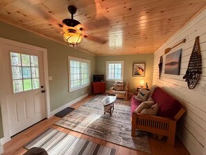 A cozy living room with a wooden ceiling, a ceiling fan, a fireplace, a sofa, a coffee table, and a TV.