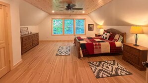Une chambre avec un plafond en bois, un lit recouvert d’une couverture à carreaux rouges et blancs, une table de chevet avec une lampe, et une fenêtre donnant sur des arbres.