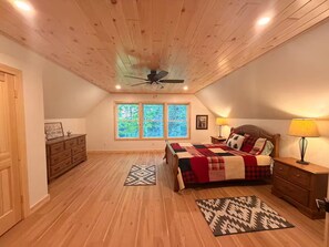 A bedroom with a wooden ceiling, a bed with a red and white checkered blanket, a nightstand with a lamp, and a window with a view of trees.