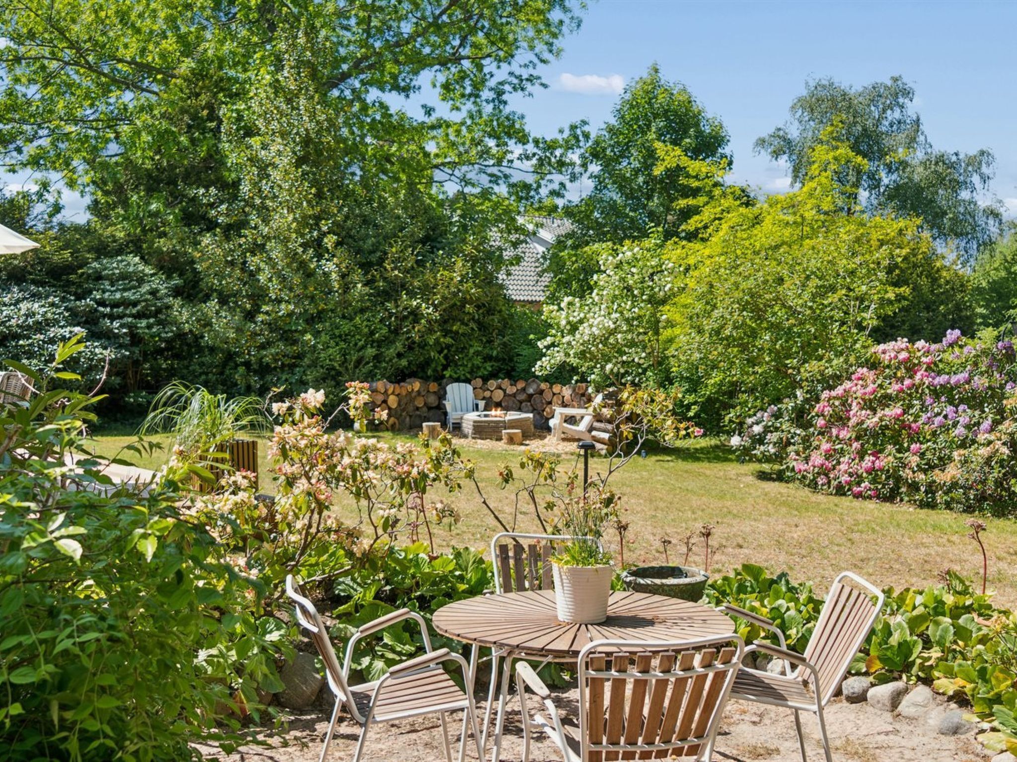A garden with a round table and chairs, surrounded by various plants and trees.