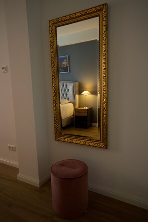 A mirror with an ornate gold frame, a tufted headboard bed, a bedside table with a lamp, and a round ottoman. - Nikolai Suite (Hamburg)