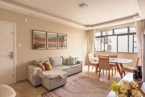 Ein Wohnzimmer mit einer Couch, einem Esstisch mit Stühlen, einem Fenster mit Vorhängen und einer Wand mit gerahmten Bildern.