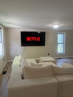 Ein Wohnzimmer mit einem großen Flachbildfernseher an der Wand, der 'NETFLIX' in roten Buchstaben anzeigt, einem beigefarbenen Ecksofa und einem Fenster mit Jalousien.