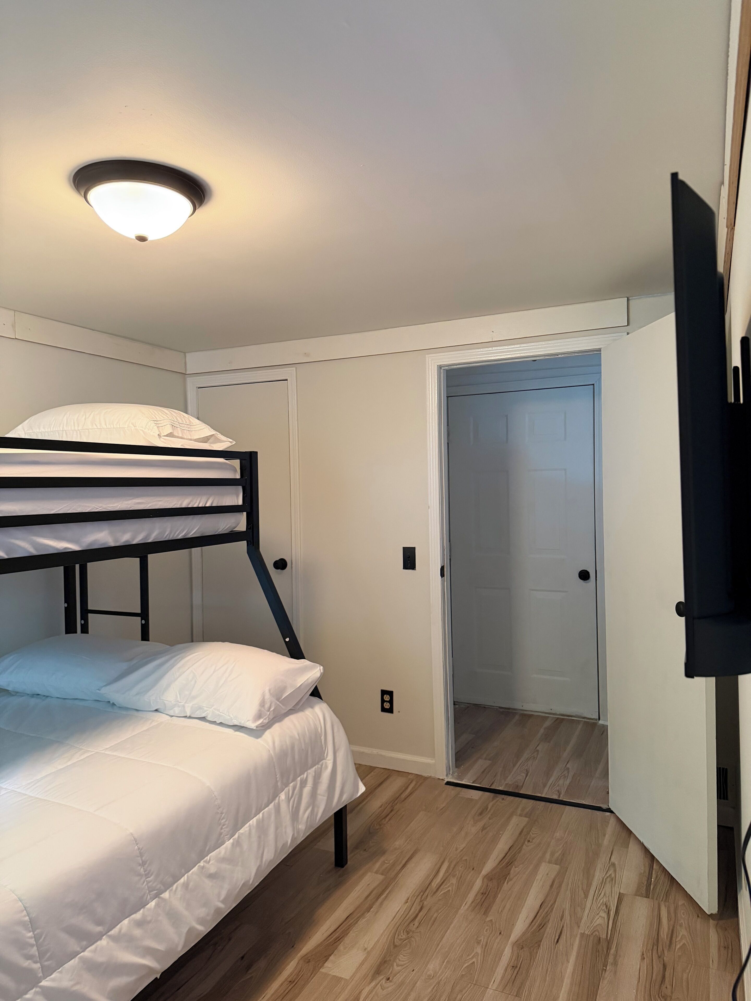 Un lit superposé avec une literie blanche, un luminaire au plafond, un plancher en bois et une porte donnant sur une autre pièce.
