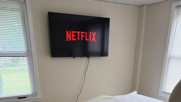 Ein Schlafzimmer mit einem Bett, einem an der Wand montierten Fernseher, der 'NETFLIX' zeigt, und Fenstern mit Jalousien.