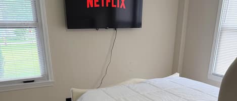 A bedroom with a bed, a wall-mounted TV displaying 'NETFLIX', and windows with blinds.