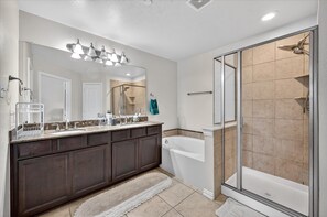 A bathroom with a double vanity, a bathtub, a shower with glass doors, and a large mirror with lights. - Modern 3 Bedroom Retreat/Open Living, Two-Car Garage & Community Pool Access (Lewisville)