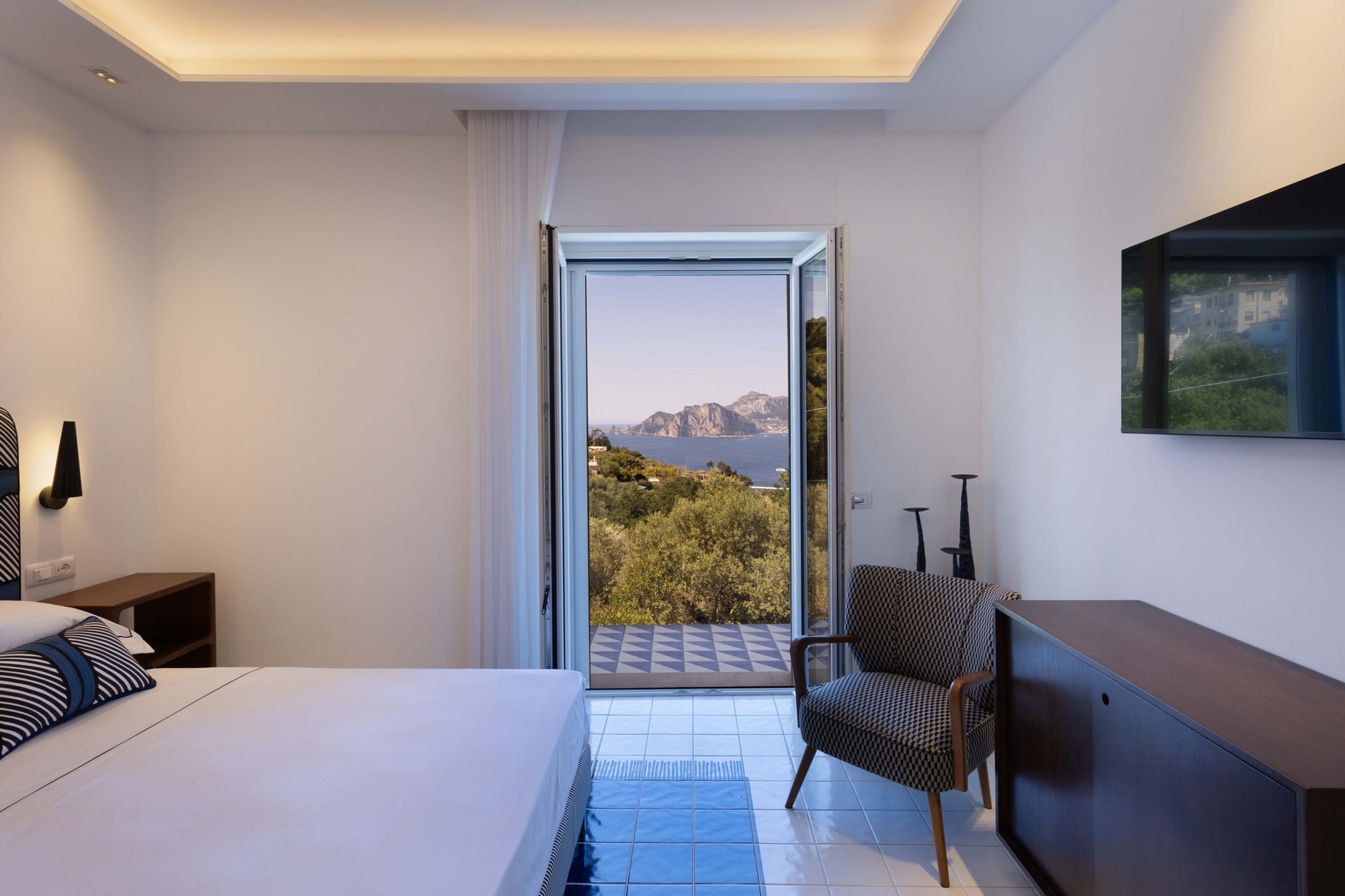 Quarto de hotel com cama, uma cadeira, escrivaninha e uma grande janela com vista para o exterior.