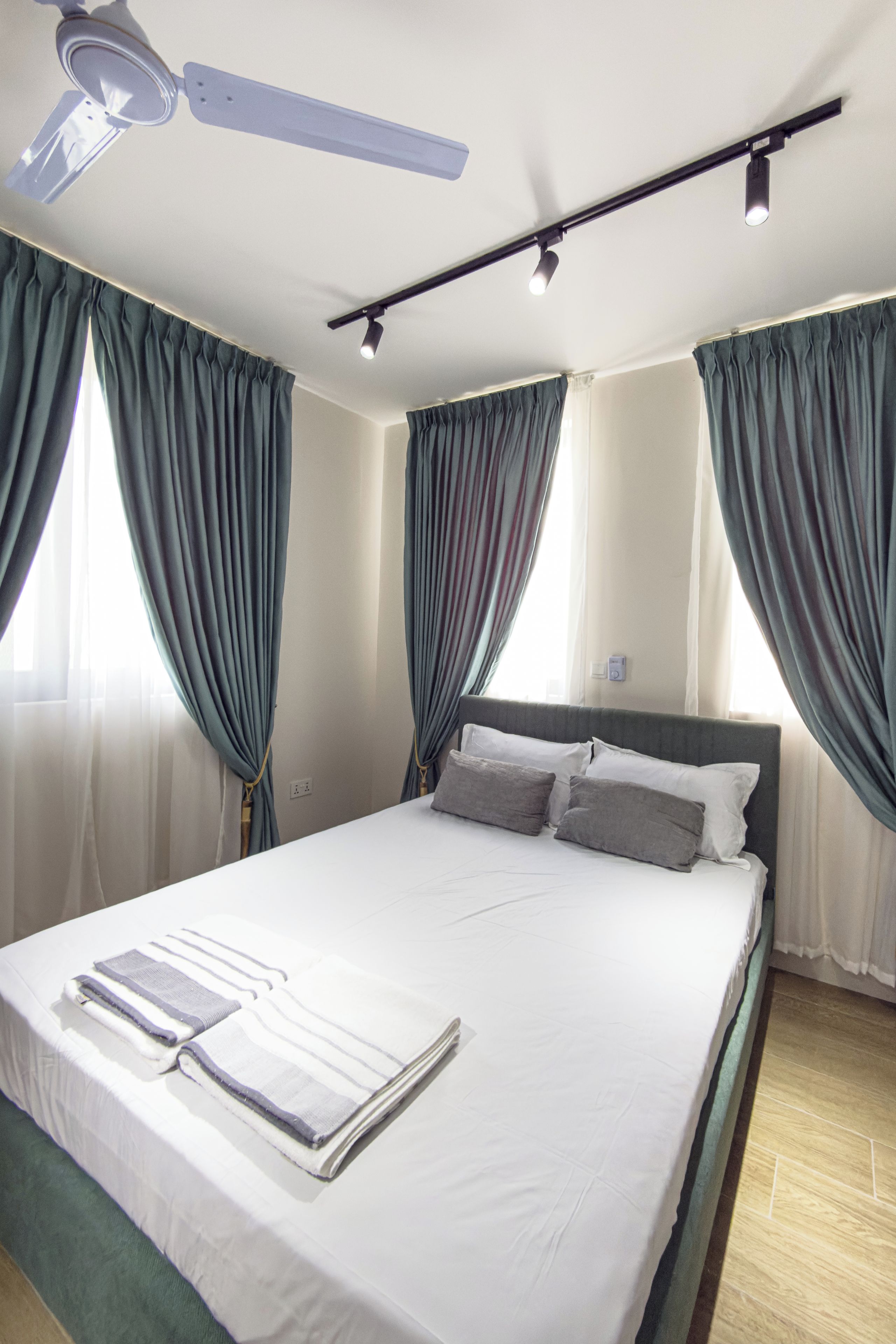 A hotel room with a large bed, two windows with curtains, a ceiling fan, and a wall-mounted light fixture.