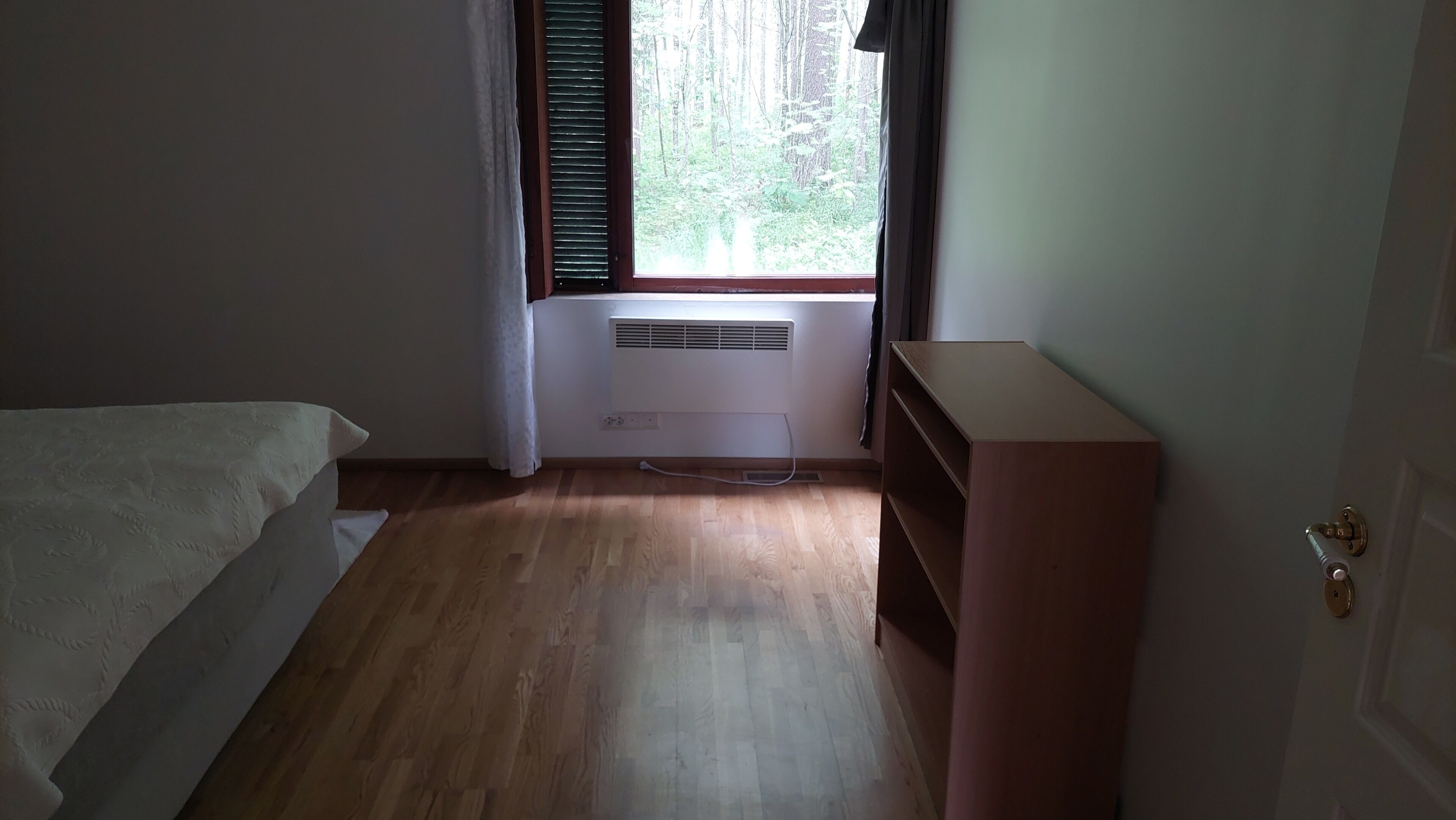 A room with a bed, a wooden floor, a window with blinds, an air conditioner, and a door.