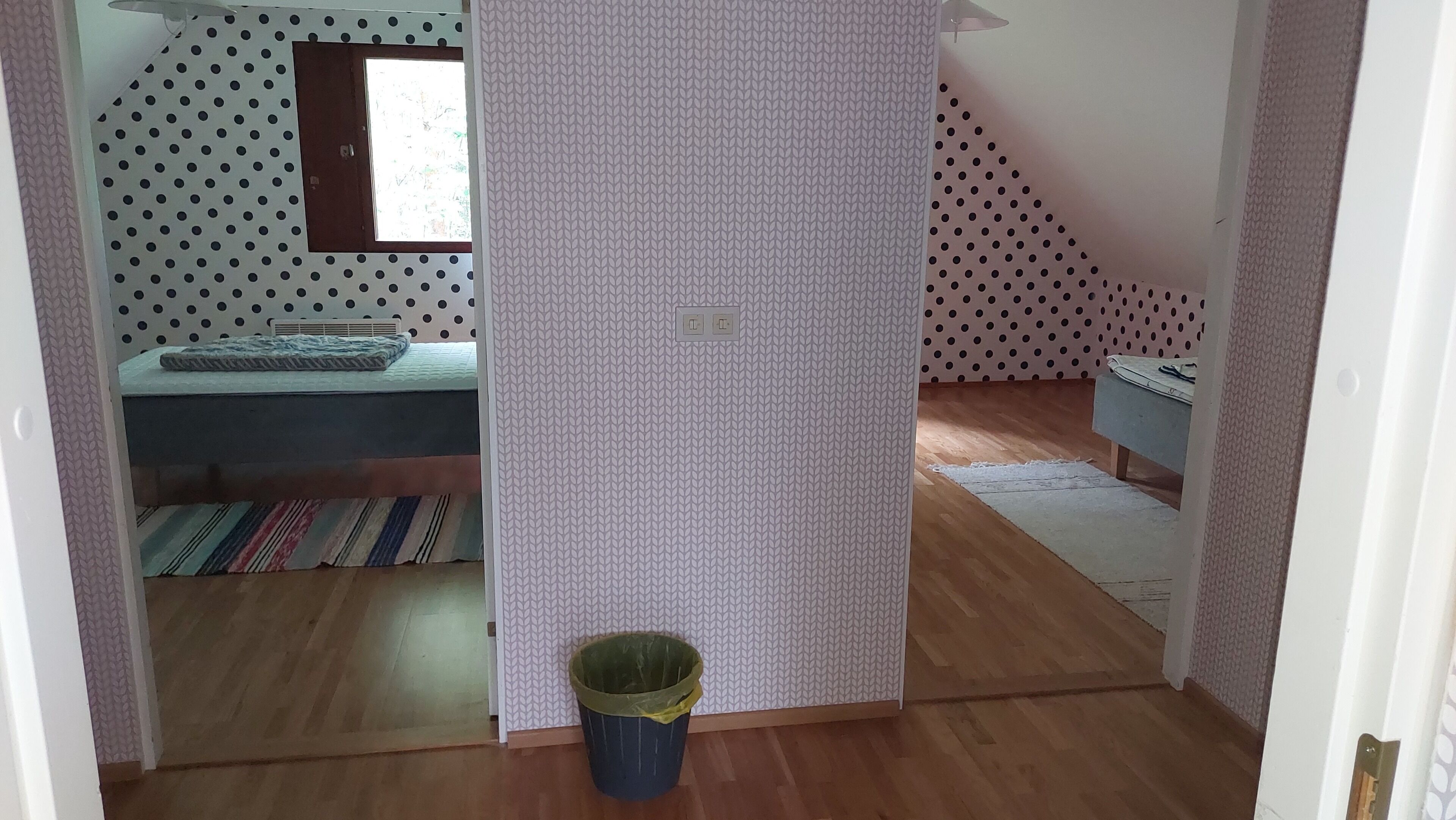 Un dormitorio con cama, ventana, pared estampada, cesto de basura y suelo de madera.