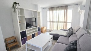 A modern living room with a grey sofa, a white coffee table, a TV on a white stand, and a window with curtains. - Large Duplex Penthouse with terrace, AC, Wifi and very spacious beds (Orpesa)