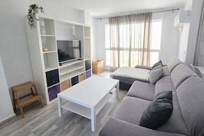 Ein modernes Wohnzimmer mit grauem Sofa, einem weißen Couchtisch, einem Fernseher auf einem weißen Schrank und einem Fenster mit Vorhängen.