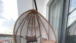 Una cadira de vusta penjant amb coixins a un balcó amb gespa artificial i vista a edificis.