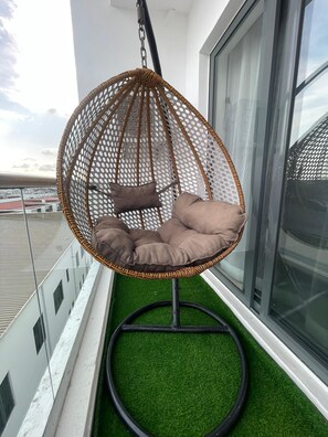 Ein Hängekorbstuhl mit Kissen auf einem Balkon mit künstlichem Rasen und Blick auf Gebäude.