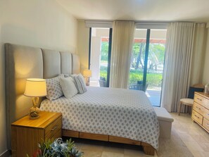 Un dormitorio con una cama grande, mesitas de noche, un aparador de madera y vistas al exterior con vegetación.