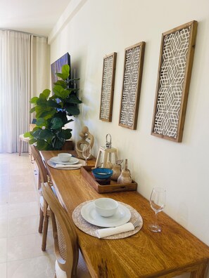 Un comedor con mesa de madera dispuesta para dos, una planta en maceta, paneles decorativos en la pared y vista al salón.