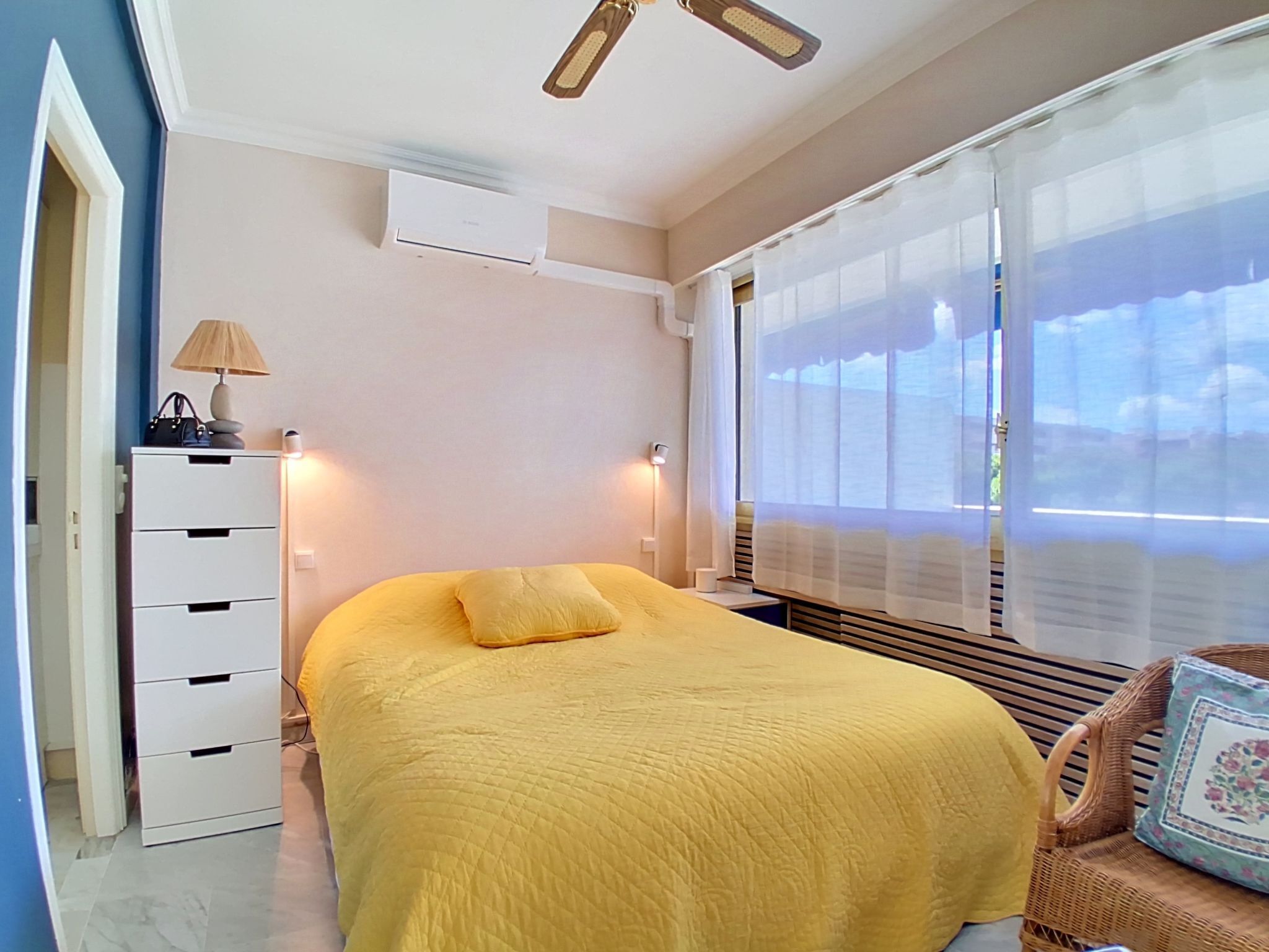 A bedroom with a bed, bedside lamp, dresser, window with sheer curtains, and a ceiling fan.