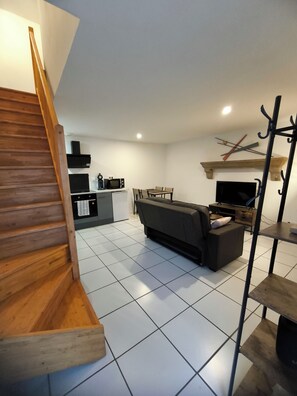 A modern living space with a wooden staircase, a sofa, a TV, and a kitchen area. - T2 apartment for 2 to 4 people (Les Epesses)