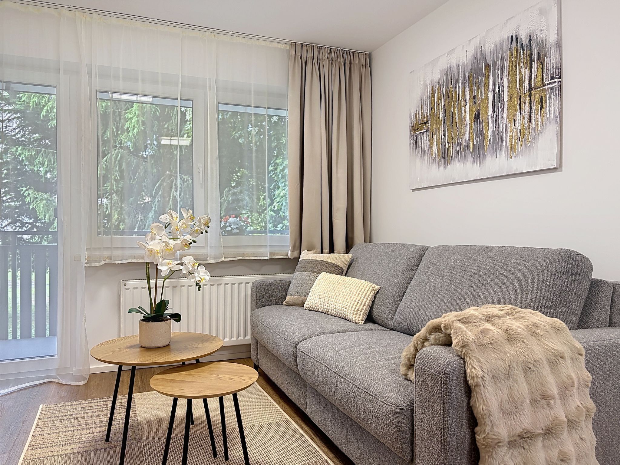 A living room with a grey sofa, a small round table, a potted plant, and a large abstract painting on the wall.