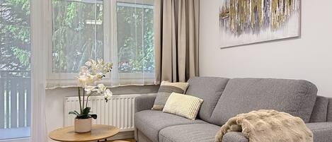 A living room with a grey sofa, a small round table, a potted plant, and a large abstract painting on the wall.
