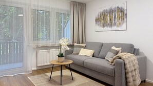 Un salon avec un canapé gris, une petite table ronde, une plante en pot et une grande fenêtre ornée de rideaux légers.