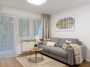 Ein Wohnzimmer mit grauem Sofa, einem kleinen runden Tisch, einem Blumentopf und einem großen Fenster mit leichtem Vorhang.