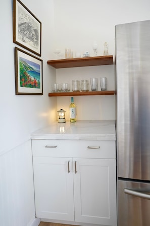 Eine Küche mit weißen Schränken, einem Edelstahlkühlschrank, einem Holzregal mit Gläsern und zwei gerahmten Bildern an der Wand.