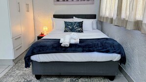 A neatly made bed with a dark blue blanket, white pillows, and a rolled towel. There is a nightstand with a lamp and a framed picture on the wall above the bed.