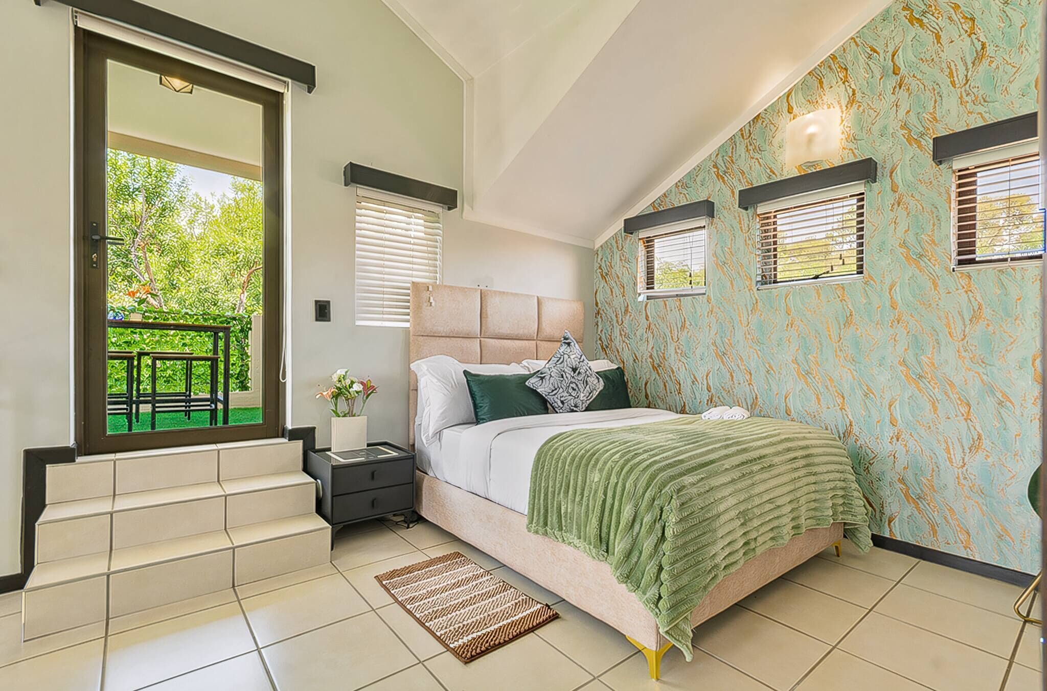 A bedroom with a bed, a nightstand, a window with blinds, a door leading to a balcony, and a patterned wall.