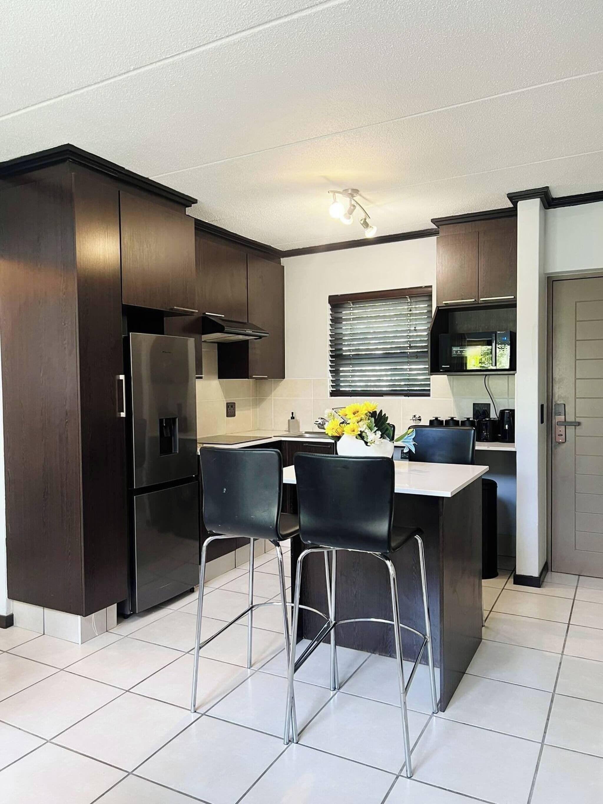A compact kitchen with dark cabinets, a stainless steel refrigerator, a microwave, and a small dining area with two chairs.