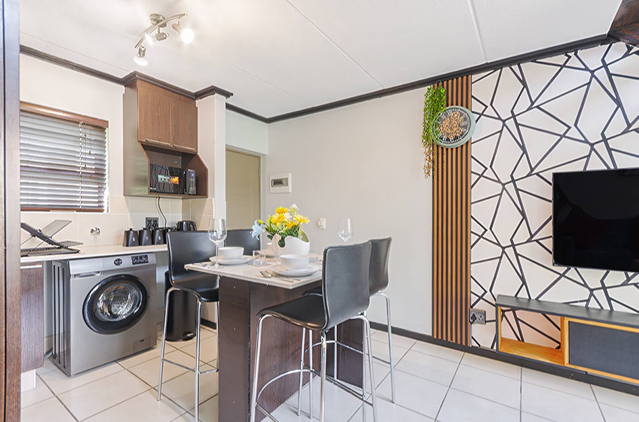 Una cocina moderna con lavadora incorporada, un comedor con mesa y sillas, y un televisor de pantalla plana montado en una pared con diseño geométrico.