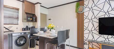Una cocina moderna con lavadora incorporada, un comedor con mesa y sillas, y un televisor de pantalla plana montado en una pared con diseño geométrico.