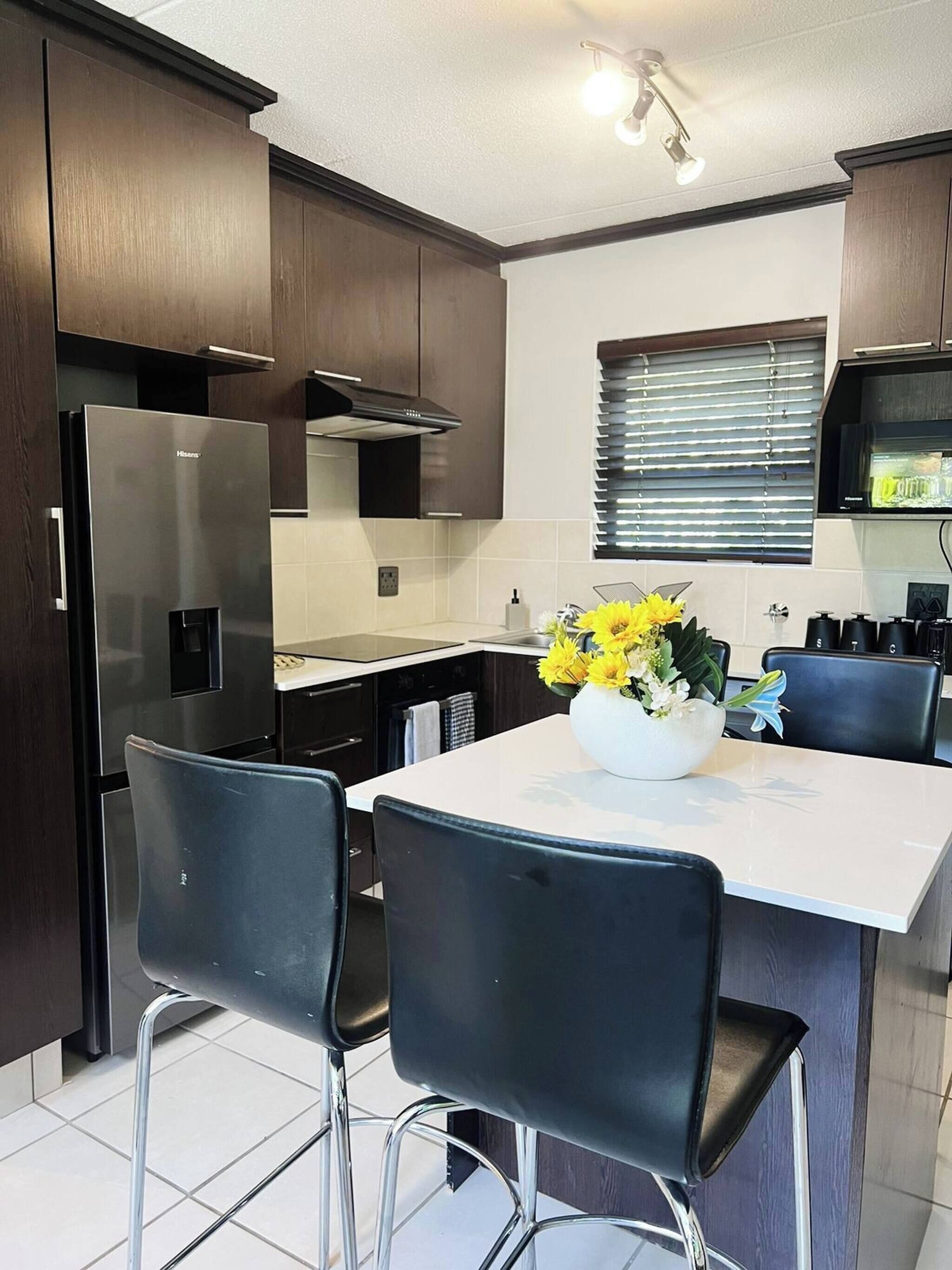 A modern kitchen with dark wooden cabinets, a stainless steel refrigerator, a white island with two chairs, and a window with blinds.