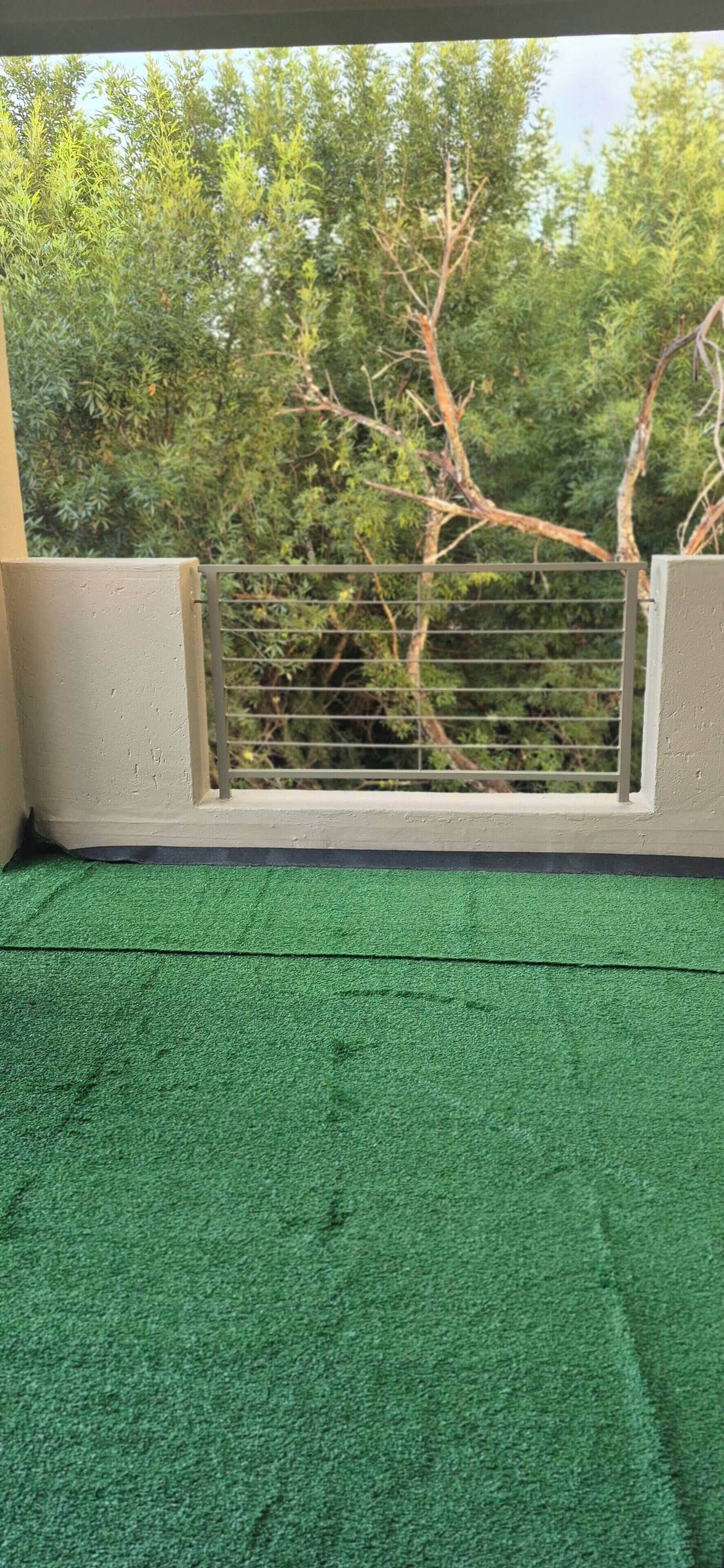 Un balcón con césped artificial verde, barandilla de metal y vistas a árboles.